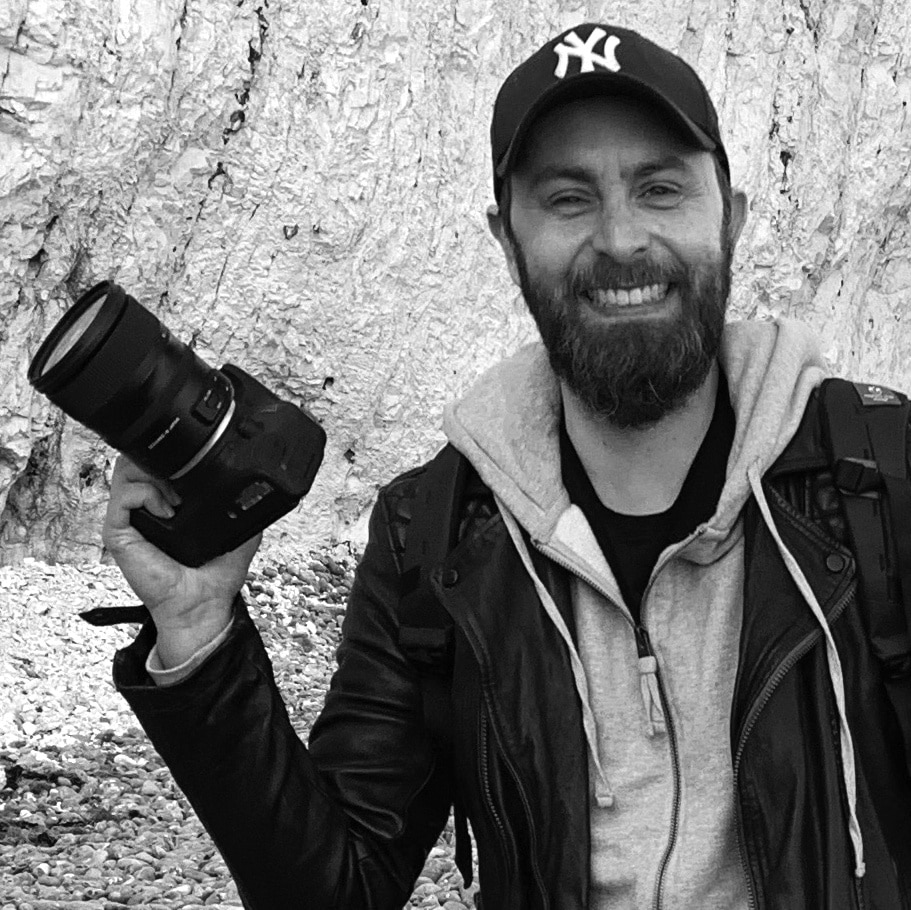 A bearded man wearing a hat, holding a camera with a smile on his face
